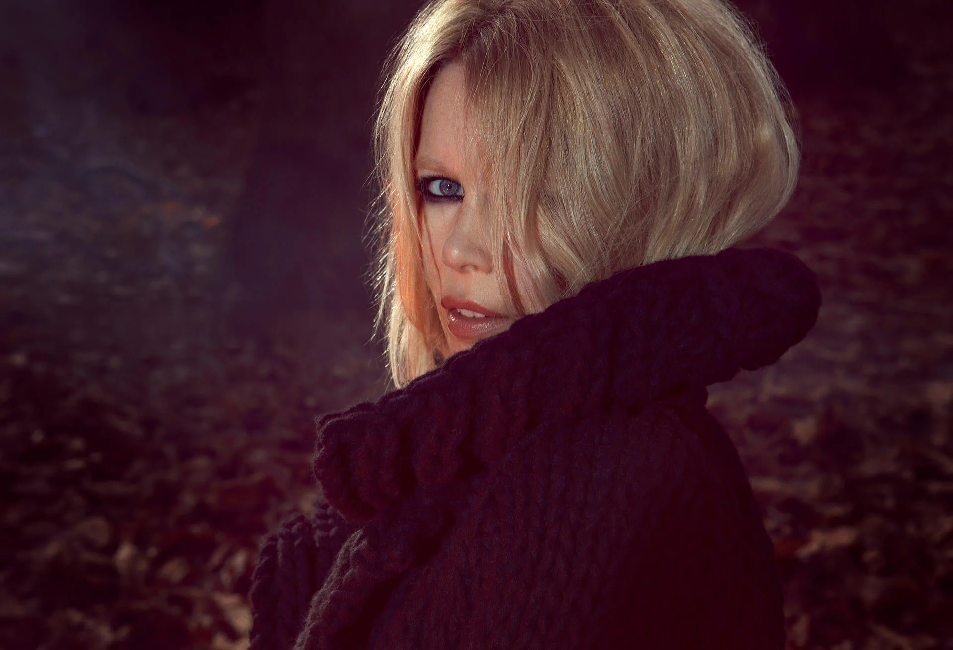 Claudia Schiffer in a dark cable-knit sweater, looking back over her shoulder in low woodland light.