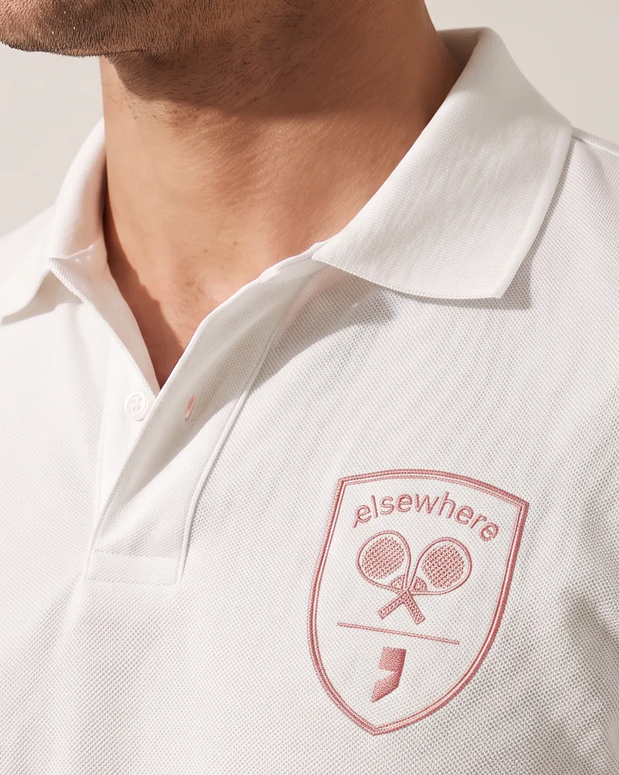 A white pique polo shirt with the Elsewhere Padel Academy crest embroidered in dusty rose at the chest.