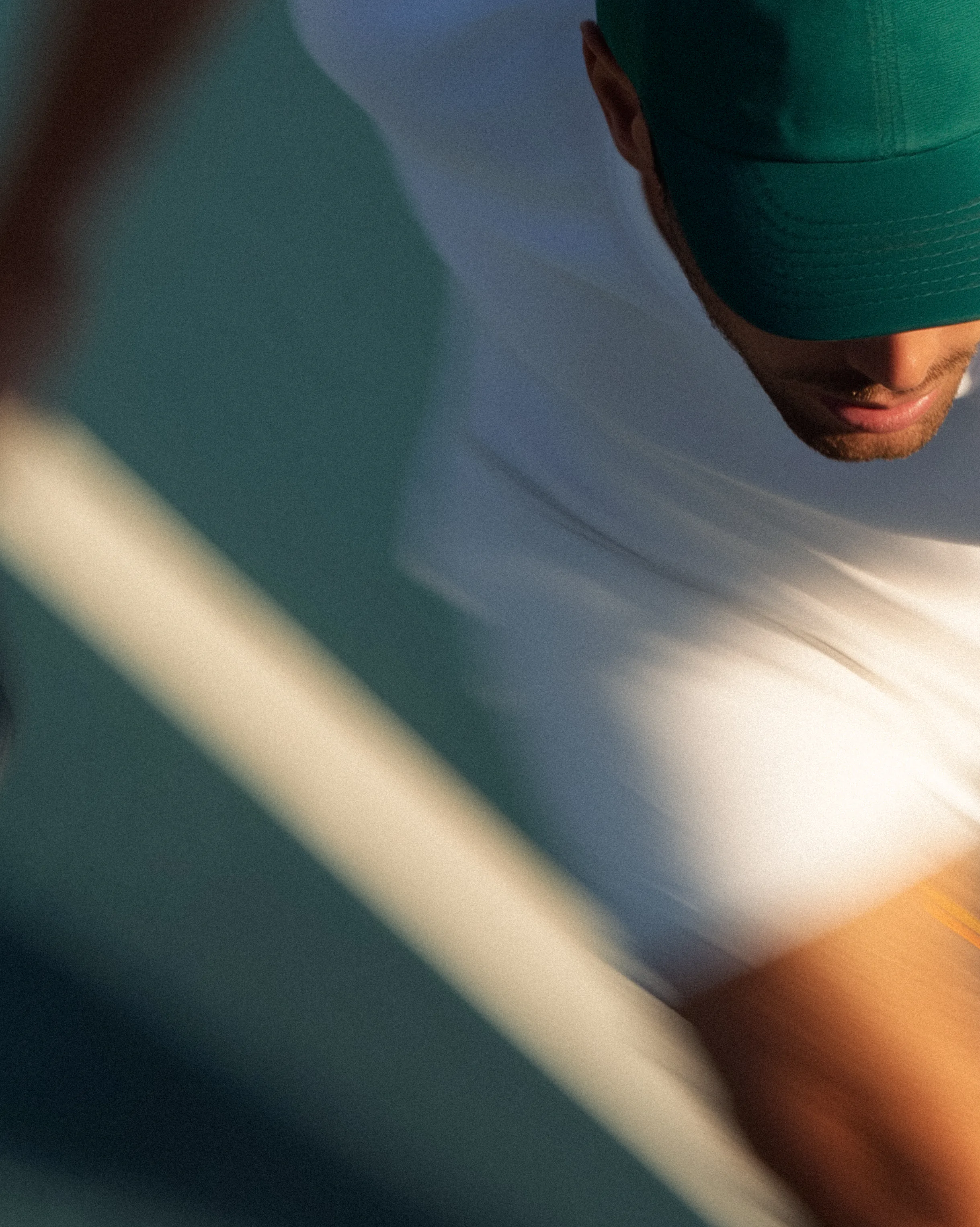 A padel player mid-stroke in white kit, seen through the court glass against a deep blue wall.