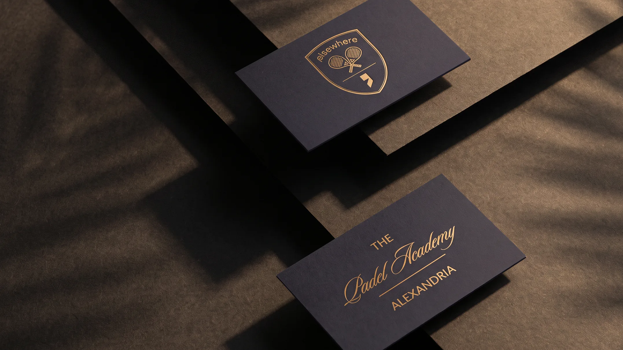 Business cards fanned on pale stone, the Elsewhere Padel Academy crest debossed on the uppermost card.