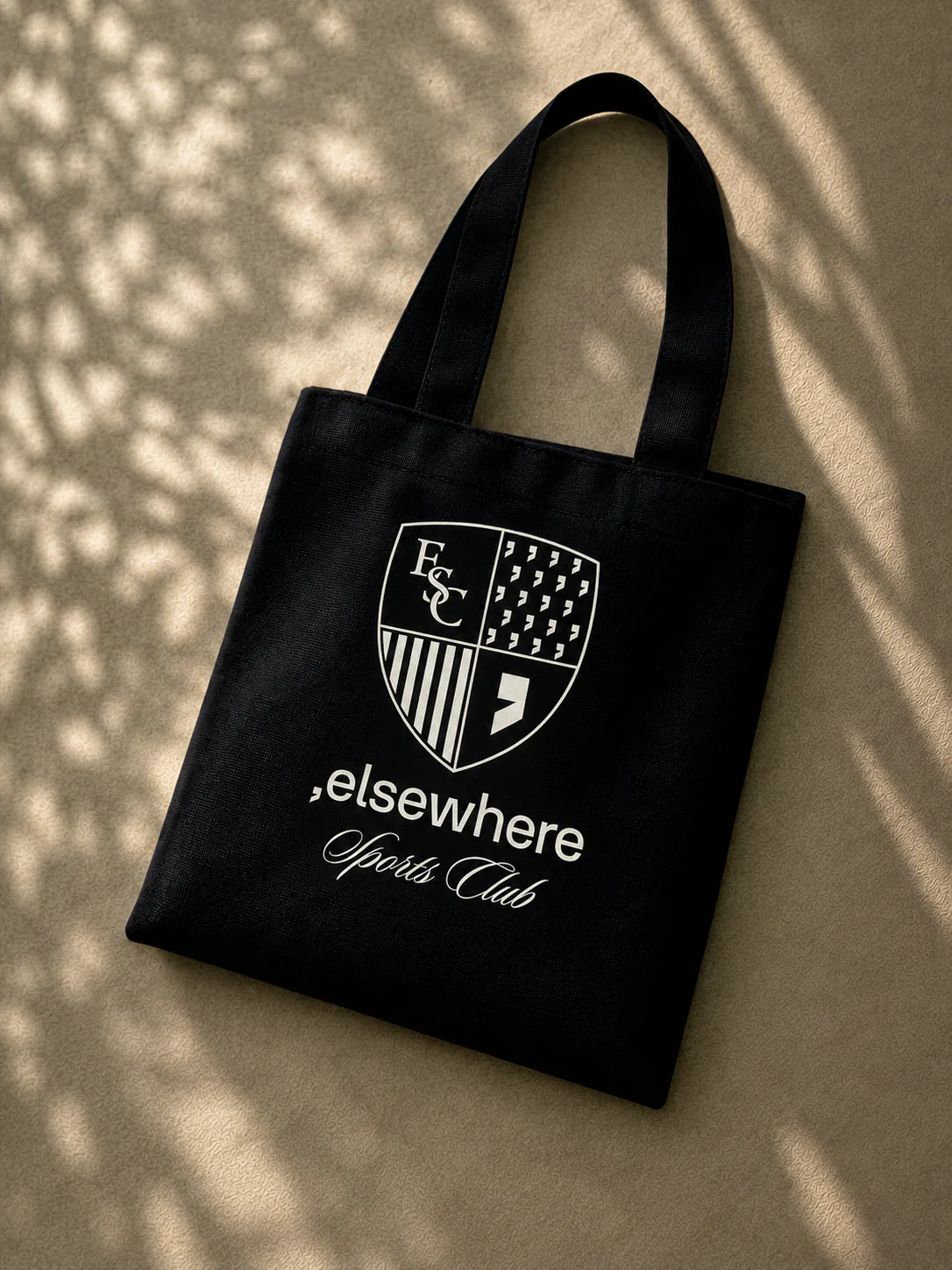 A black canvas tote printed with the Elsewhere crest and wordmark, on warm sand under dappled leaf shadow.