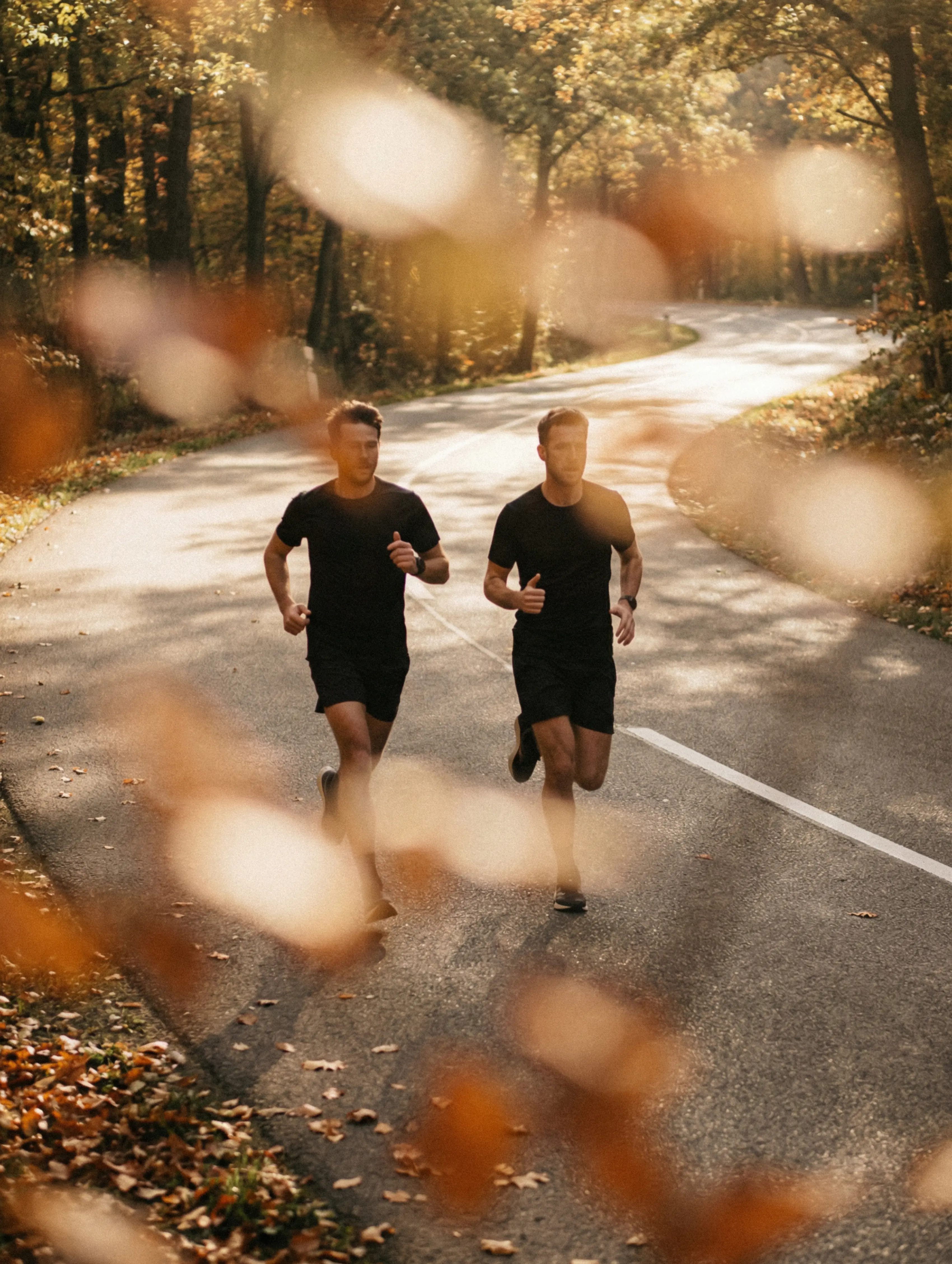 Two runners on a sunlit forest road in autumn, seen through blurred amber leaves.
