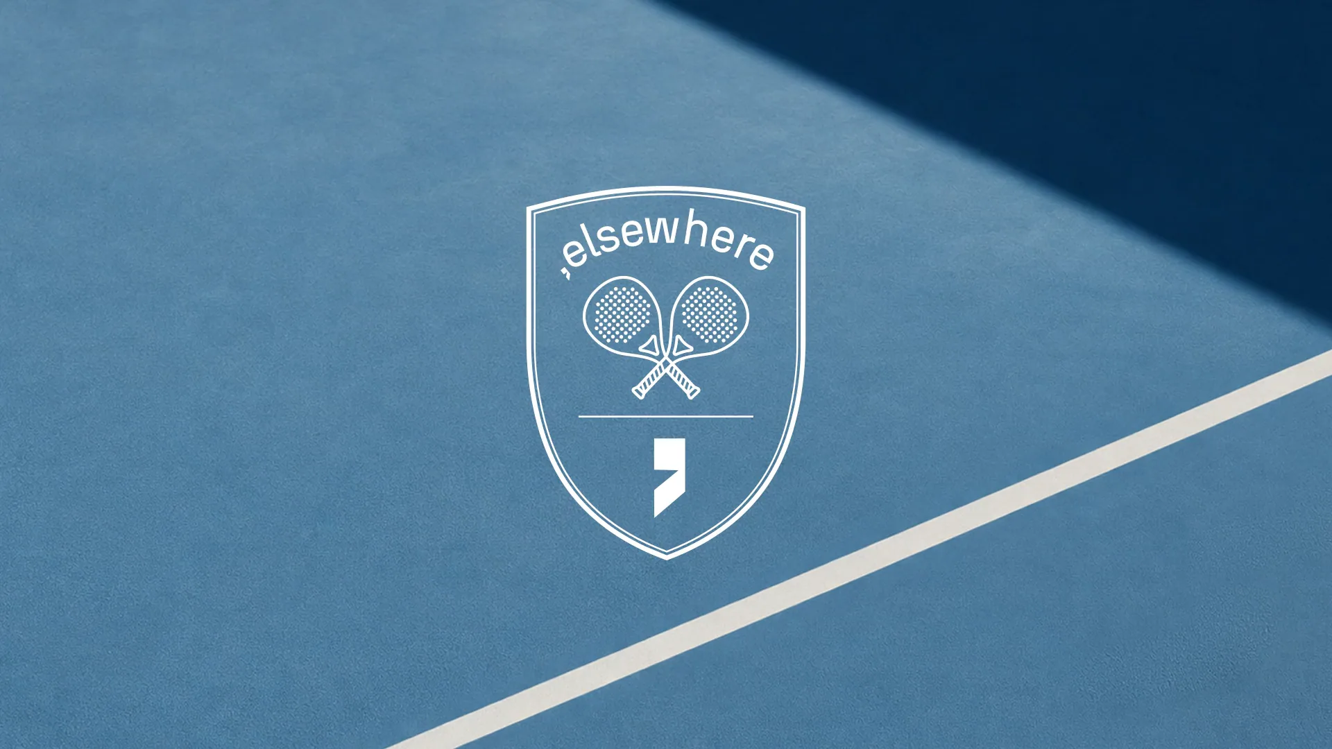 A blue padel court in low sun, the white Elsewhere crest centred, a yellow ball rolling in to rest beside the line.