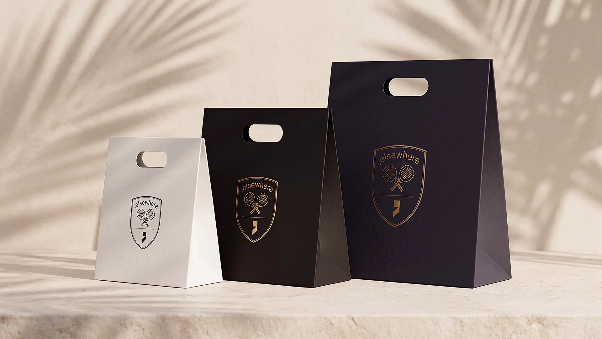 Three Elsewhere carrier bags in white, black and navy on a stone ledge, palm shadows drifting on the wall behind.
