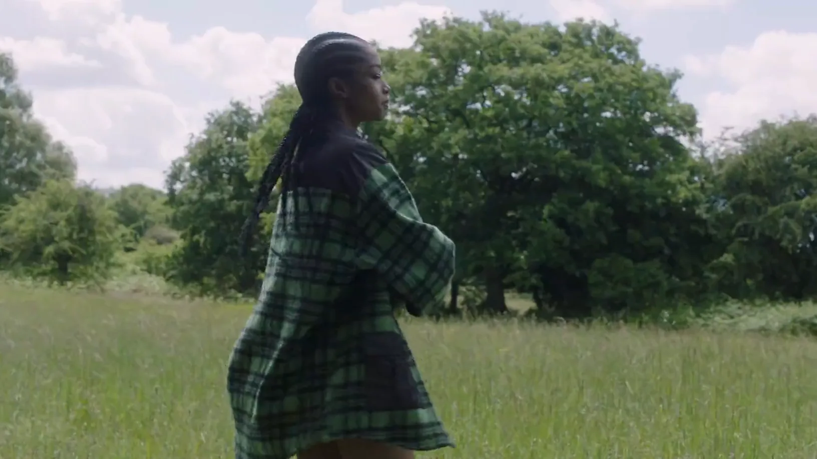 A model in a green check coat turning in a summer meadow, arms raised against cloud.