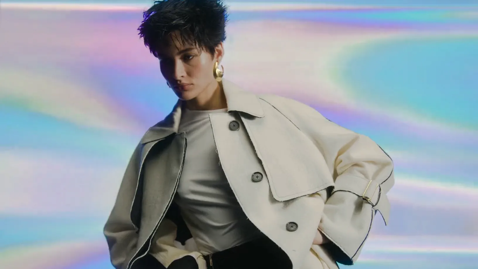 A model in a cream coat against an iridescent ground, turning later in a one-shoulder dress.