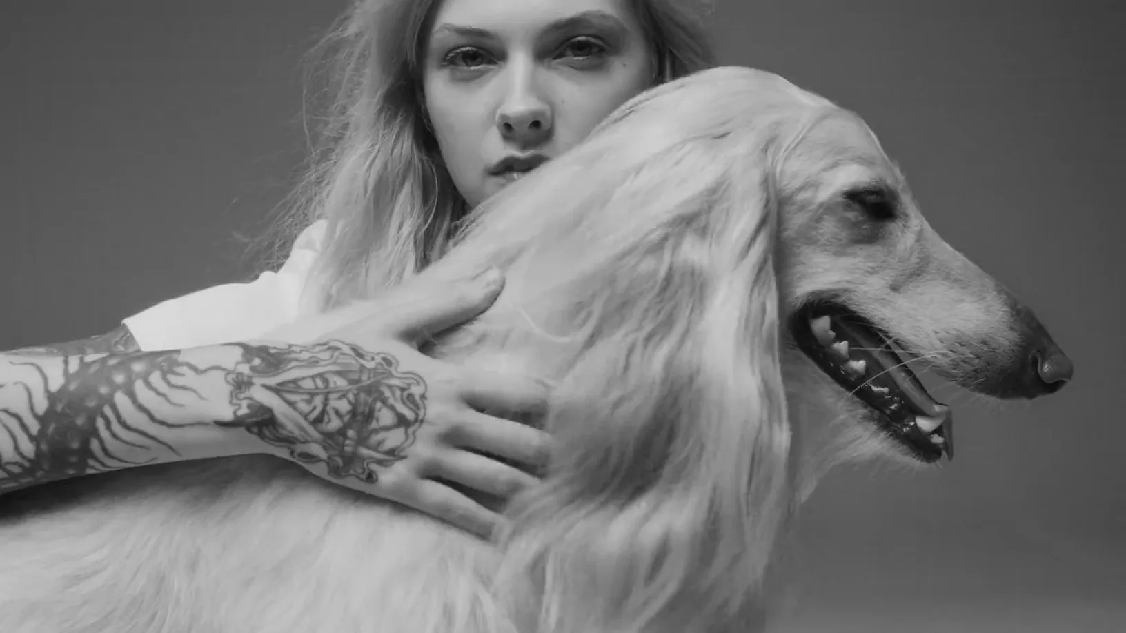 A fair-haired woman holding a long-coated dog in a grey studio, in black and white.