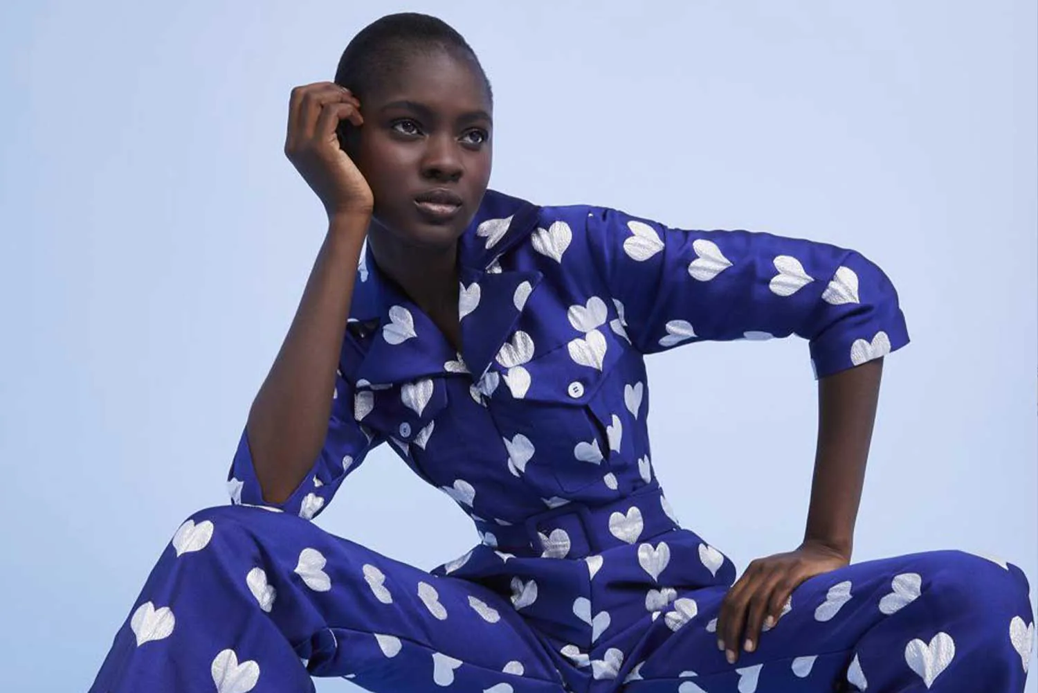 A model seated in a blue heart-print silk shirt and trousers, against pale blue.