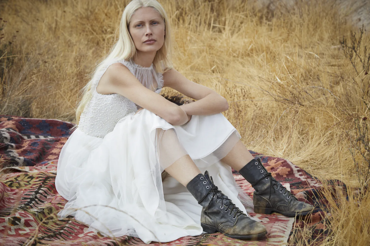 A model seated on a patterned rug in dry grass, in a white dress and black lace-up boots.