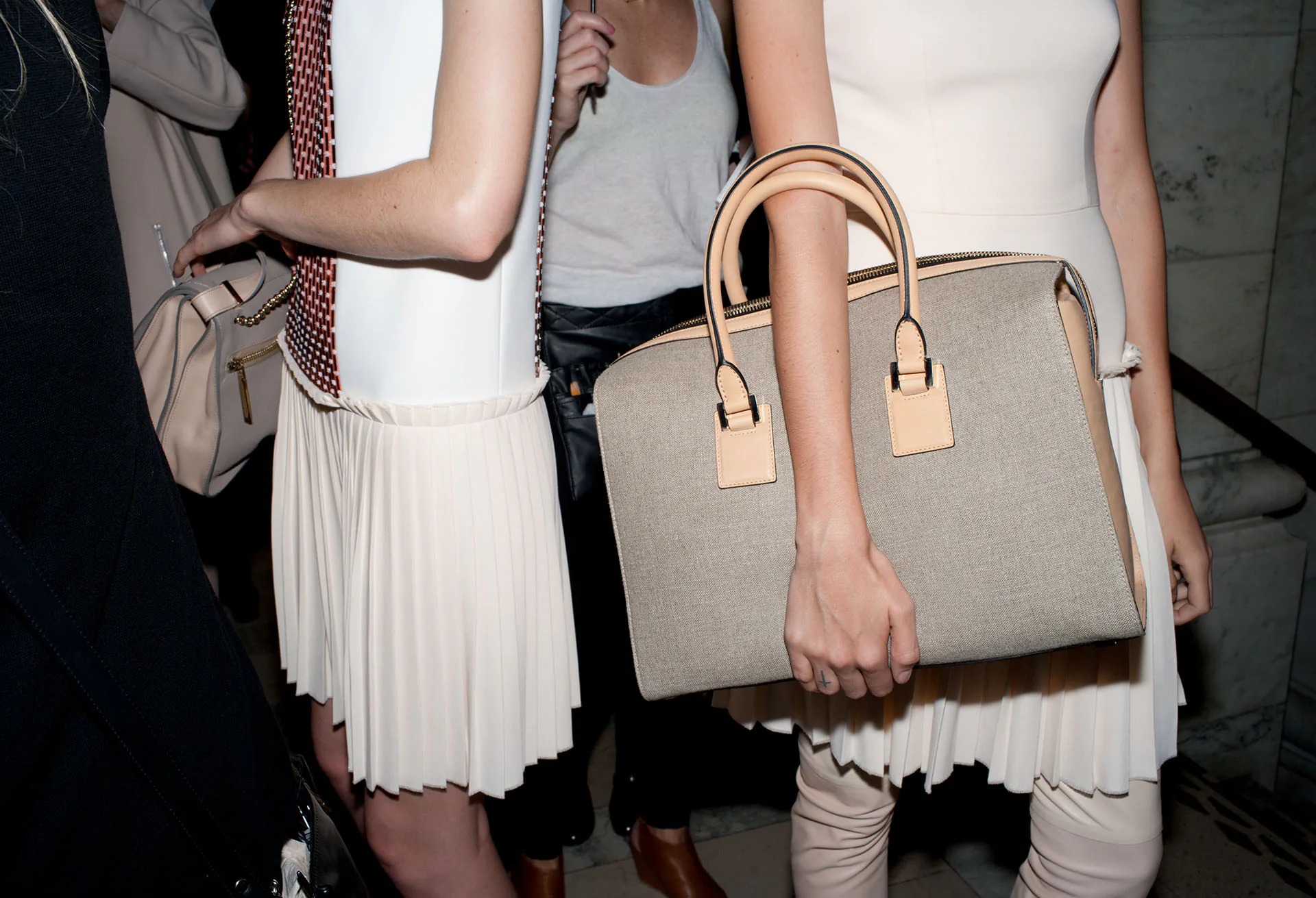 A model in a pleated white skirt holding a large structured tote with rolled handles.